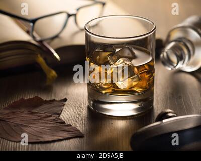 Hintergrundbeleuchtetes Glas Whisky auf den Felsen auf einem Holztisch, mit Notizbuch, Stift und Lesebrille Stockfoto