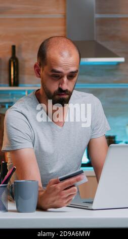 Junger Mann, der spät in der Nacht online shoppen und in der Küche mit Kreditkarte auf dem Laptop sitzt. Mitarbeiter, der von zu Hause aus eine Zahlungstransaktion auf einem digitalen Notebook macht, das mit dem Internet verbunden ist Stockfoto