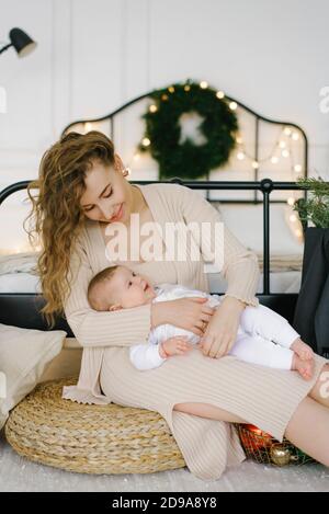 Mama und Baby kuscheln sich auf dem Hintergrund eines Bettes und eines Weihnachtsbaums, Konzepterferien, Neujahr und Weihnachten in der Familie Stockfoto