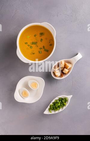 Bouillon in weißer Suppenschüssel, gehackte Frühlingszwiebeln oder Frühlingszwiebeln, Wachteleier und Croutons auf grauem Steingrund. Tabellenansicht von oben. Vertikale Orientatio Stockfoto