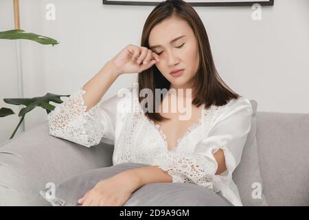 Asiatische Frauen in einem Nachtkleid und tragen weiße Satin-Robe, die nachts auf dem grauen Sofa im Wohnzimmer sitzt, EIN Mädchen in SeidenNachtwäsche sc Stockfoto