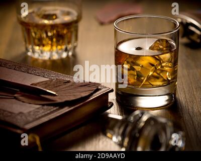 Hintergrundbeleuchtetes Glas Whisky auf den Felsen auf einem Holztisch, mit Notizbuch, Stift und einem zweiten Glas Stockfoto