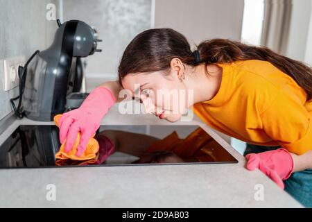 Hausarbeit. Porträt einer Hausfrau in Gummihandschuhen, fleißig wischen den Induktionsherd mit einem Lappen. Seitenansicht. Stockfoto