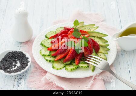 Veganer Salat aus Erdbeere, Gurke und Avocado mit Mohn, Öldressing und Minze. Frische, gesunde, diätetische Lebensmittel. Auf einem weißen Teller und auf einer blu Stockfoto