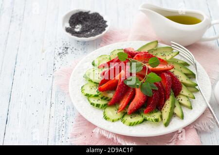 Veganer Salat aus Erdbeere, Gurke und Avocado mit Mohn, Öldressing und Minze. Frische, gesunde, diätetische Lebensmittel. Auf einem weißen Teller und auf einer blu Stockfoto