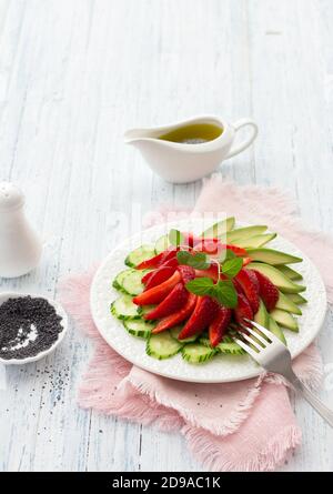 Veganer Salat aus Erdbeere, Gurke und Avocado mit Mohn, Öldressing und Minze. Frische, gesunde, diätetische Lebensmittel. Auf einem weißen Teller und auf einer blu Stockfoto