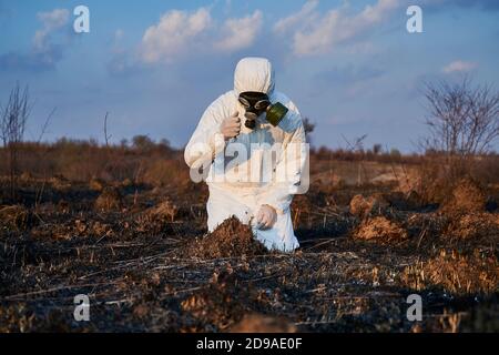 Männlicher Ökologe in Schutzanzug und Gasmaske Ausgießen Boden aus der Hand, während Forschungsarbeit auf verbranntem Feld, Analyse Zustand des Bodens. Konzept von Ökologie, Forschung und verbrannter Erde. Stockfoto