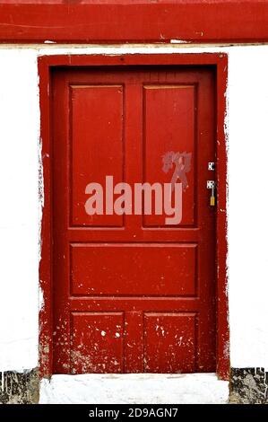 Eine verschlossene Farbe spritzte rote Holztür in einem rustikalen weißen Steingebäude. Stockfoto