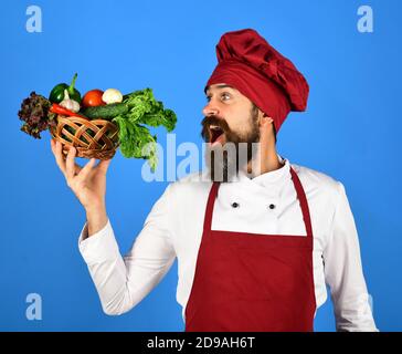 Kochen Sie mit fröhlichem Gesicht in burgunderroten Uniform hält Gemüse in Weidenschale. Der Küchenchef hält Salat, Tomaten, Pfeffer und Pilze bereit. Vegetarisches Diät-Konzept. Mann mit Bart auf blauem Hintergrund. Stockfoto