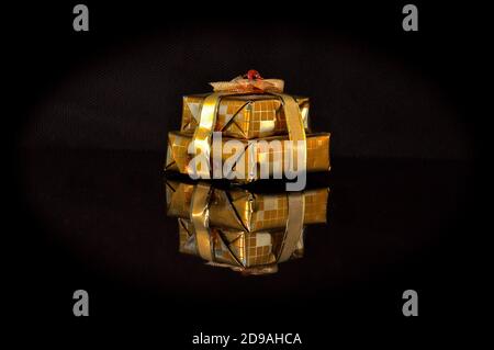 Kleine cuboid Pakete Geschenk in Goldfolie Geschenkpapier und glänzendem Goldband verpackt, isoliert auf einer schwarzen reflektierenden Oberfläche, zeigt eine umgekehrte mirro