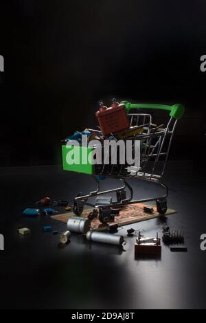 Metallwagen Warenkorb, gefüllt mit Funkkomponenten, steht auf einer elektronischen Platine. Foto in dunkler Hintergrundbeleuchtung. Stockfoto