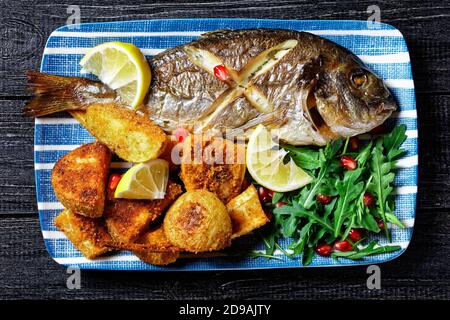 Gerösteter dorado-Fisch mit gebackenen Kartoffeln in Paniermehl serviert Auf einem Teller mit einem Zitronenkeil und frisch wild Rucola Salat mit Granatapfelkernen auf Stockfoto