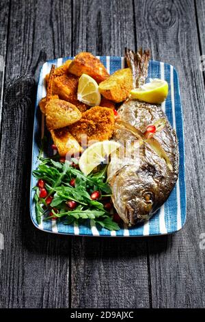 Gerösteter dorado-Fisch mit gebackenen Kartoffeln in Paniermehl serviert Auf einem Teller mit einem Zitronenkeil und frisch wild Rucola Salat mit Granatapfelkernen auf Stockfoto