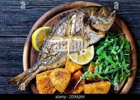 Ganze geröstete dorado Fisch mit Haut auf gebackenen Kartoffeln beschichtet In Paniermehl serviert auf einem hölzernen Schneidebrett mit Zitrone Keil und frische Rucola oder wild Stockfoto