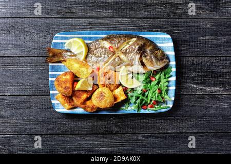 Gerösteter dorado-Fisch mit gebackenen Kartoffeln in Paniermehl serviert Auf einem Teller mit einem Zitronenkeil und frisch wild Rucola Salat mit Granatapfelkernen auf Stockfoto