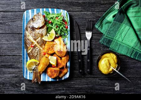 Gerösteter dorado-Fisch mit gebackenen Kartoffeln in Paniermehl serviert Auf einem Teller mit einem Zitronenkeil und frisch wild Rucola Salat mit Granatapfelkernen auf Stockfoto