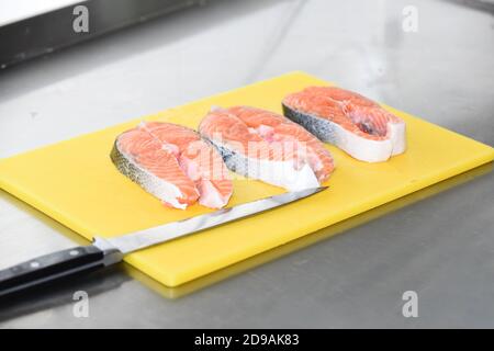 Schneidebrett in gelber Farbe mit frischem Lachs und Messer. Rohe Lachssteaks mit Messer auf Schneidebrett. Tisch mit Lachssteaks und Messer auf verschwommenem Hintergrund. Meeresfrüchtekonzept. Stockfoto