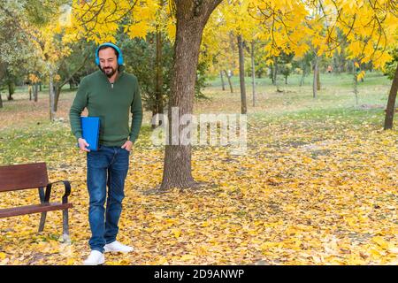 Junger bärtiger Hipster Mann steht mit blauen kabellosen Kopfhörern und Laptop unter seinem Arm Vorbereitung auf die Arbeit in einem Park Im Herbst mit gefallenen Baum gehen Stockfoto