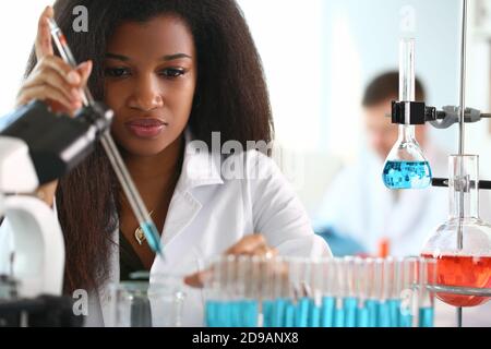 Eine afrikanische Frau im Labor mit Mikroskop und Flüssigkeiten in Kolben, die mit ihnen arbeiten Stockfoto