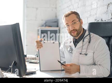 Der Arzt zeigt mit einem Stift auf die Zwischenablage. Er schaut auf die Kamera und zeigt die Testergebnisse. Stockfoto