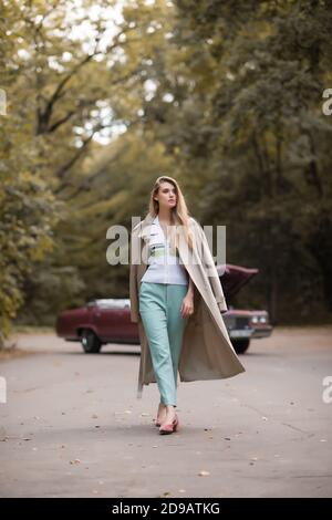 Stilvolle Frau im Umhang weg beim Gehen auf der Straße schauen In der Nähe gebrochen Retro-Auto auf verschwommenem Hintergrund Stockfoto