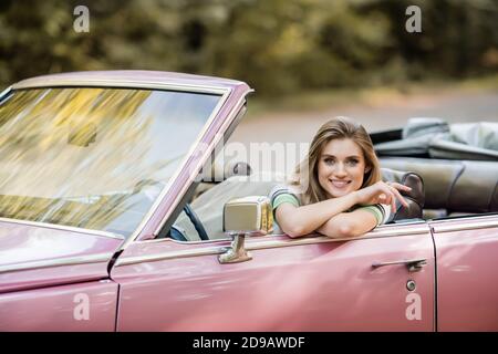 Glückliche junge Frau, die im Retro-Cabriolet sitzt und lächelt Bei der Kamera Stockfoto