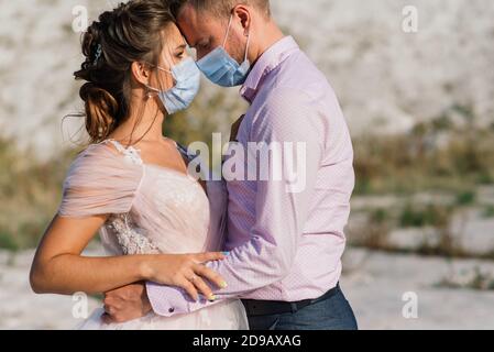Junge liebende Paar in medizinischen Masken im Park während der Quarantäne an ihrem Hochzeitstag zu Fuß. Stockfoto