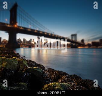 Felsige Küste des East River mit Blick auf die verschwommene Skyline von Manhattan und die Manhattan Bridge bei Nacht. Stockfoto