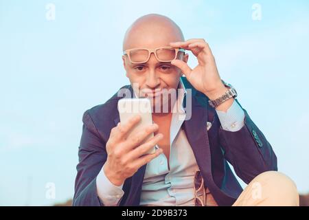 Nahaufnahme Porträt Kopfschuss Mann mittleren Alters mit Brille skeptisch Blick auf den Handy-Bildschirm sitzen im Freien blauen Himmel auf dem Hintergrund. Negatives fa Stockfoto