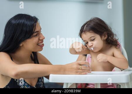 Eine lächelnde, ethnische hispanische Mutter füttert einem zweijährigen Mädchen, das auf einem Hochstuhl in einem Zimmer zu Hause sitzt, Joghurt. Stockfoto