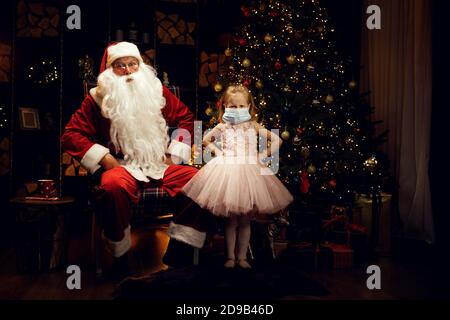 Santa Claus und kleine böse kleine Mädchen machen sie tragen eine medizinische Maske in Weihnachtsferien und Pandemie. Stockfoto