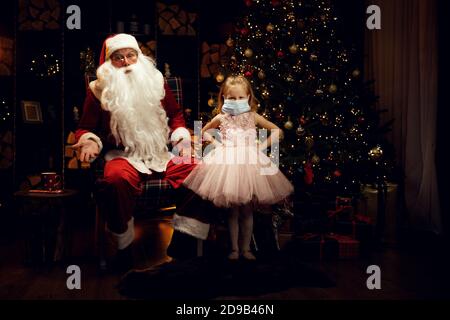 Santa Claus und kleine böse kleine Mädchen machen sie tragen eine medizinische Maske in Weihnachtsferien und Pandemie. Stockfoto