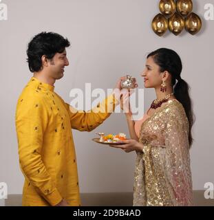 Gut aussehende Ehemann Durchführung Karwa Chauth Traditionen des Angebots Wasser zu ihrer fastender Frau in traditioneller Kleidung. Stockfoto