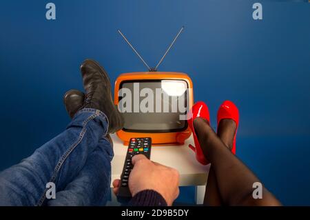 Ein Ehepaar vor einem kleinen alten Fernseher wirft ihre Füße in Schuhe auf den Tisch und schaltet ihn mit der Fernbedienung ein. Stockfoto
