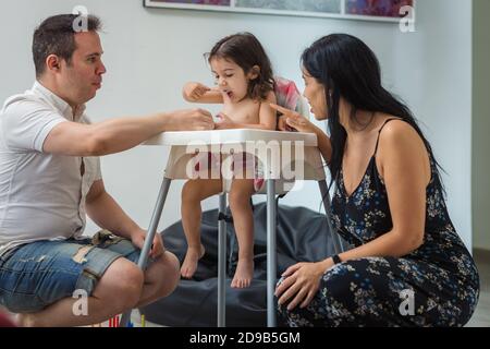 Eine lächelnde, ethnische hispanische Mutter und kaukasischer Vater füttert einem zweijährigen Mädchen, das auf einem Hochstuhl in einem Zimmer zu Hause sitzt, Joghurt. Stockfoto