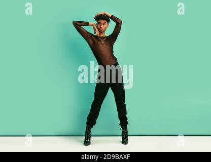 In voller Länge von einem Homosexuell Mann in schwarz Outfit. Geschlecht fließender Mann trägt schwarze Jeans, schwarzes Netzhemd und High Heels posiert vor blauem Hintergrund. Stockfoto