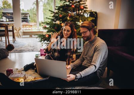 Paar mit Laptop für Videoanrufe ihre Familie an einem Weihnachtstag. Mann und Frau mit einem Videoanruf mit geschmückten Weihnachtsbaum im Hintergrund. Stockfoto