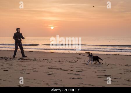 Ein Mann geht mit seinem jungen Hund in den frühen Morgenstunden auf lange Leine am Strand von Bournemouth in Dorset, während die Sonne gegen einen orangefarbenen Januarhimmel über dem Meer aufgeht. 23. Januar 2015. Foto: Neil Turner Stockfoto