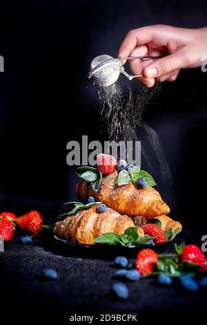 Zerbröckelnder Effekt. Der Koch streut das Croissant mit Puderzucker. Croissants auf dunklem Hintergrund. Croissant mit Beeren. Französisches Frühstück mit Stockfoto