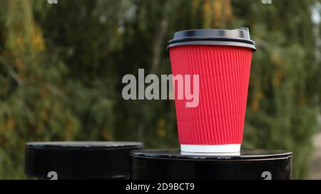 Kaffee in einem Papier Einweg-Öko-Glas der roten Farbe mit einem schwarzen Kunststoffdeckel vor dem Hintergrund eines grünen Parks. Selektiver Fokus. Nahaufnahme. Stockfoto