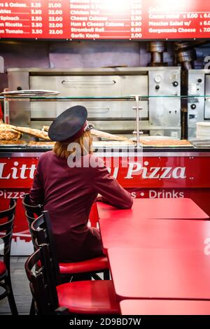 NEW YORK, USA - 02. Mai 2016: Junge Frau in einem burgunderroten Anzug und Mütze sitzt an einem roten Tisch in der Pizzeria und erwartet Pizza Stockfoto