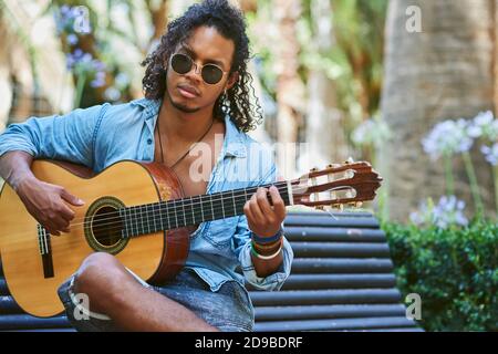 Junger Musiker mit klassischer Gitarre, der im Schatten einiger Bäume in einem Park spielt. Stockfoto