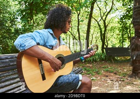 Junger Musiker mit klassischer Gitarre, der im Schatten einiger Bäume in einem Park spielt. Stockfoto