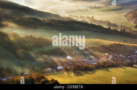 Luftaufnahme des Dorfes im Nebel mit goldenen Sonnenstrahlen bei Sonnenaufgang im Herbst. Schöne ländliche Landschaft mit Straße, Gebäuden, nebligen bunten Bäumen. Slovaki Stockfoto