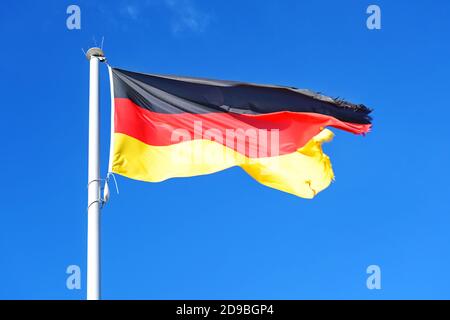 Deutsche Flagge mit Streifen in schwarz, rot, Gold auf einem Mast, flattert mit ausgefranster Naht im Wind gegen einen blauen Himmel, Kopierraum, Bewegungsunschärfe Stockfoto