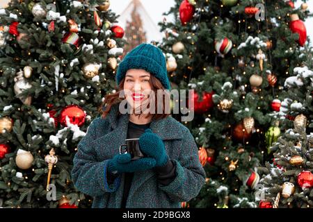 Schöne Brünette Frau Holding Tasse heißen Kakao oder Tee mit Weihnachtsbaum auf dem Hintergrund. 2021. Verbringen Sie Nw Jahresurlaub Mit Freude Und Familie Stockfoto