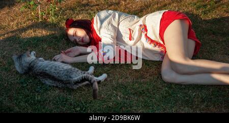 Junge Frau mit scharlachroten Dreadlocks in Nationaltracht auf dem Gras liegen und mit der Katze spielen. Außenportrait Stockfoto