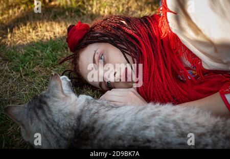 Junge Frau mit scharlachroten Dreadlocks in Nationaltracht auf dem Gras liegen und mit der Katze spielen. Außenportrait Stockfoto