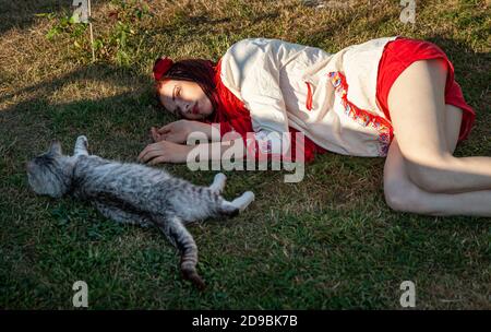 Junge Frau mit scharlachroten Dreadlocks in Nationaltracht auf dem Gras liegen und mit der Katze spielen. Außenportrait Stockfoto