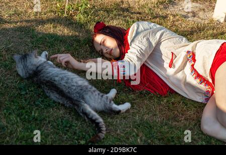 Junge Frau mit scharlachroten Dreadlocks in Nationaltracht auf dem Gras liegen und mit der Katze spielen. Außenportrait Stockfoto